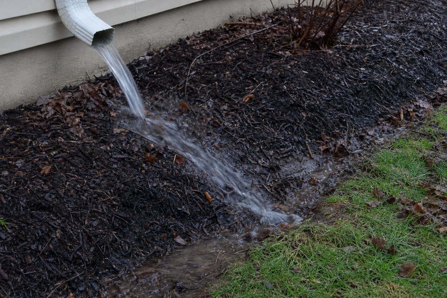 Downspout outlet splashing onto a mulch bed with visible washout toward the lawn